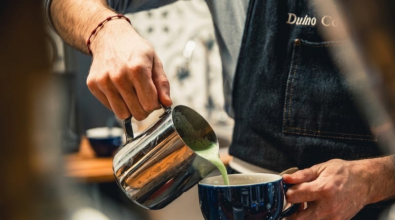 A barista in a denim apron pours green matcha latte from a metal pitcher into a blue mug.