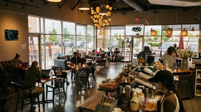 A bustling coffee shop interior with customers working on laptops and a barista preparing drinks.