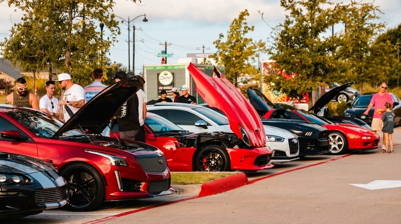 People gather around luxury cars with open hoods at an outdoor car show.