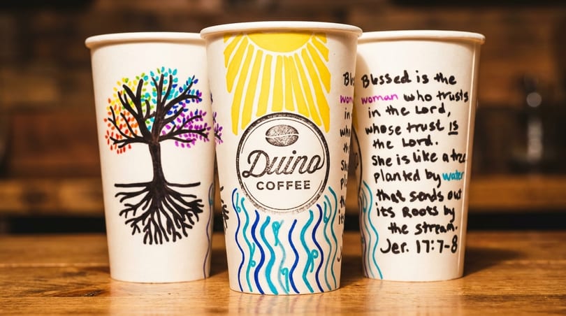 Three Duino Coffee cups with hand-drawn designs, including a tree, sun, and scripture.