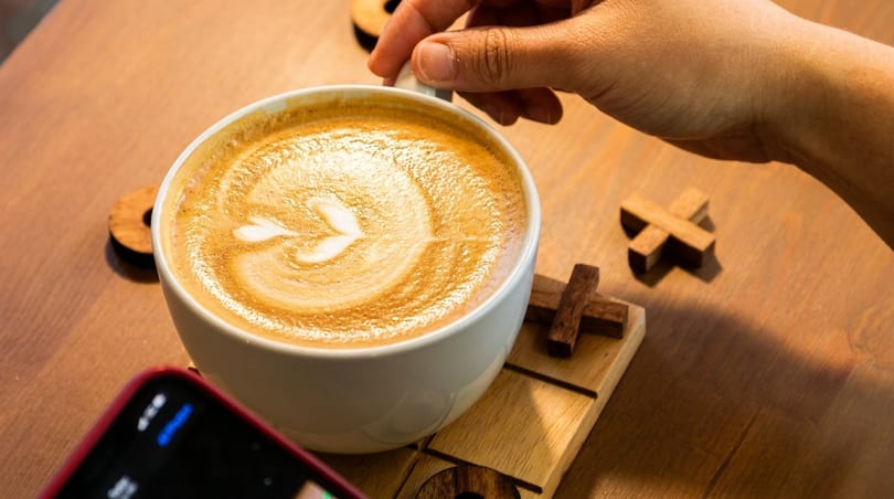 Hand holding a latte with heart art, phone, and wooden tic-tac-toe pieces on a table.