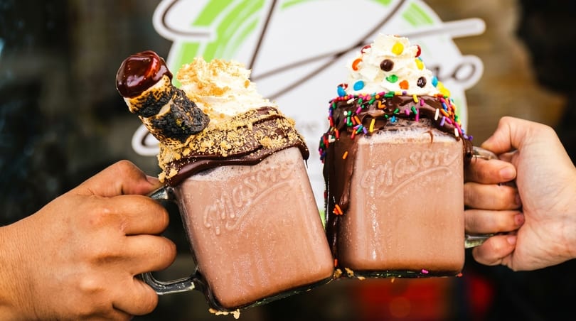 Two hands holding up chocolate milkshakes in mason jars, one with a toasted marshmallow, the other with sprinkles.