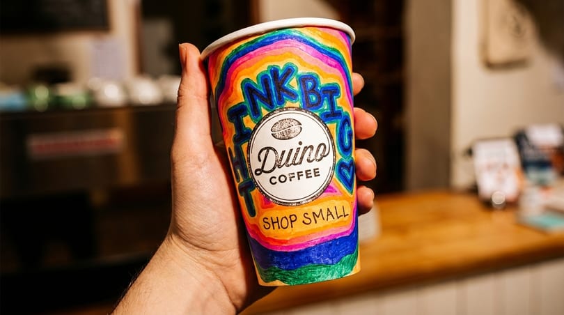 Hand holding a colorful Duino Coffee cup with "Think Big Shop Small" written on it.