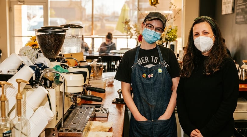 Two masked baristas stand behind a coffee bar with espresso machines and coffee bean grinders.